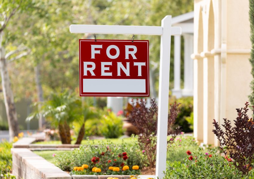 A red and white 'for rent' sign hands off a white post in front of a rental property's lawn.